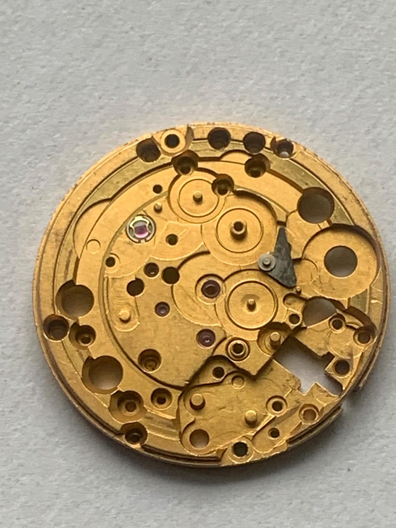 May include: Close-up of a gold-colored watch movement with intricate gears, jewels, and components. The circular mechanism features numerous holes and small details, showcasing the internal workings of a timepiece. The image highlights the precision and complexity of the watch's internal structure.