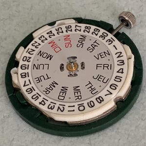May include: A watch movement with a white dial featuring the days of the week in English and numbers 1-31. The dial is surrounded by a green plastic ring and has a silver winding stem.