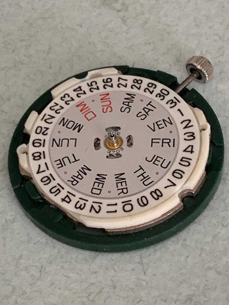 May include: A watch movement with a white dial featuring the days of the week in English and numbers 1-31. The dial is surrounded by a green plastic ring and has a silver winding stem.