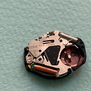 May include: Detailed view of a watch movement, showcasing various metal parts. Features include a copper coil, a black triangular element, and a red-hued circular piece. The text "SEIKO TIME CORP." is present on the mechanism.