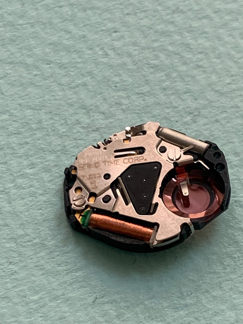 May include: Detailed view of a watch movement, showcasing various metal parts. Features include a copper coil, a black triangular element, and a red-hued circular piece. The text "SEIKO TIME CORP." is present on the mechanism.