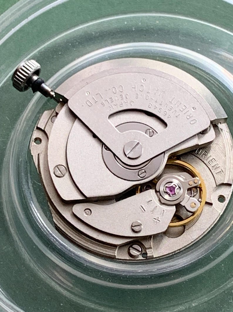 May include: Detailed view of a watch mechanism, showcasing intricate gears, screws, and a winding stem. The text "ORIENT WATCH CO. LTD" and "JAPAN" are present. The movement is silver-toned with a small, purple jewel.