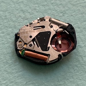 May include: A detailed view of a watch movement, featuring various metal parts, a copper coil, and a red crystal. The text "E&C TIME CORP" is visible. The movement is set against a light blue background.