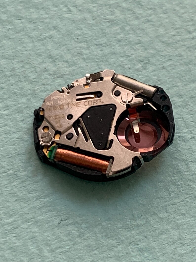 May include: A detailed view of a watch movement, featuring various metal parts, a copper coil, and a red crystal. The text "E&C TIME CORP" is visible. The movement is set against a light blue background.