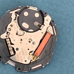 May include: Close-up of a Citizen watch movement, showcasing intricate internal components. The mechanism features a silver-colored plate with various gears, a copper-colored coil, and the text "Citizen Watch Co. 110 Jewels Japan". The watch movement is set against a blue background.