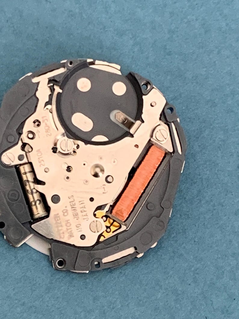 May include: Close-up of a Citizen watch movement, showcasing intricate internal components. The mechanism features a silver-colored plate with various gears, a copper-colored coil, and the text "Citizen Watch Co. 110 Jewels Japan". The watch movement is set against a blue background.