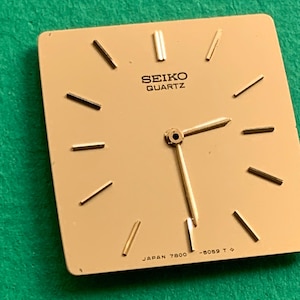 May include: A square, champagne-coloured Seiko Quartz watch dial featuring gold-tone baton hour markers and two gold-tone hands. The text SEIKO QUARTZ is printed in black near the top, with JAPAN 7800 -5059 T printed in black at the bottom.