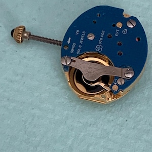 May include: A close-up of a watch movement with a blue circuit board and gold-colored components. The watch movement has the text "SWISS SIX 6 JEWELS" and "978.002" printed on it. A small screw and a gold-colored crown are also visible.