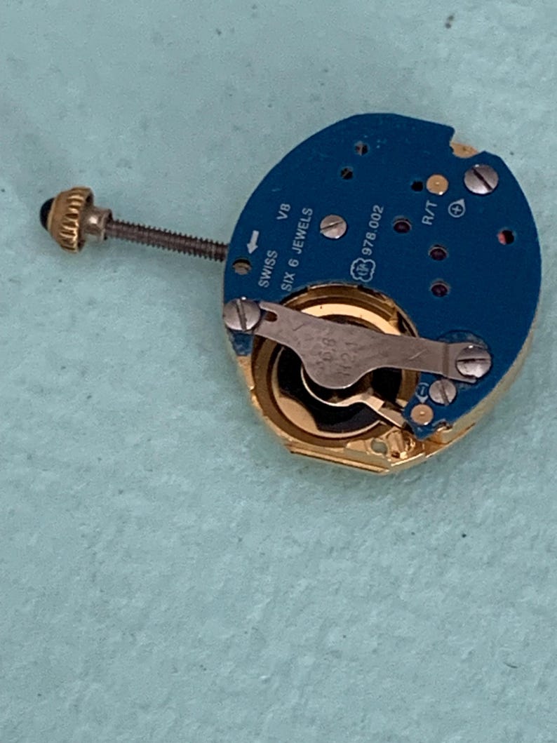 May include: A close-up of a watch movement with a blue circuit board and gold-colored components. The watch movement has the text "SWISS SIX 6 JEWELS" and "978.002" printed on it. A small screw and a gold-colored crown are also visible.