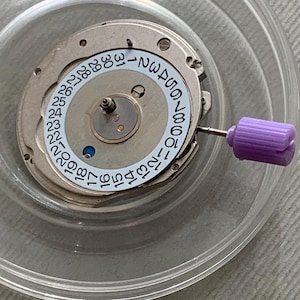 May include: A watch movement with a white date dial featuring black numbers from 1 to 31. The movement is silver-toned with small screws and a purple winding stem. The components are inside a clear plastic container.