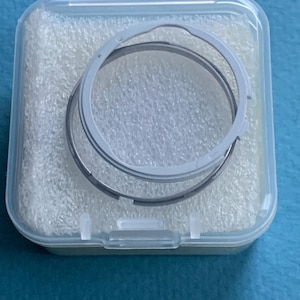 May include: A transparent plastic case holding two circular metal rings. The rings are grey and silver, and look like parts of a mechanism. The case is filled with white foam padding.