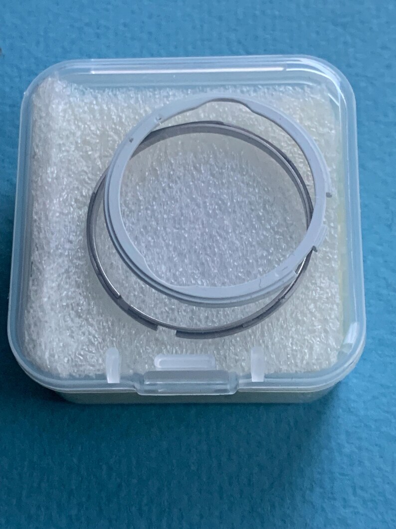 May include: A transparent plastic case holding two circular metal rings. The rings are grey and silver, and look like parts of a mechanism. The case is filled with white foam padding.