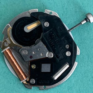 May include: Close-up of a watch movement with a black and silver design. The watch mechanism includes a battery, a copper coil, and the word "ORIENT". The watch movement is labeled "JAPAN".