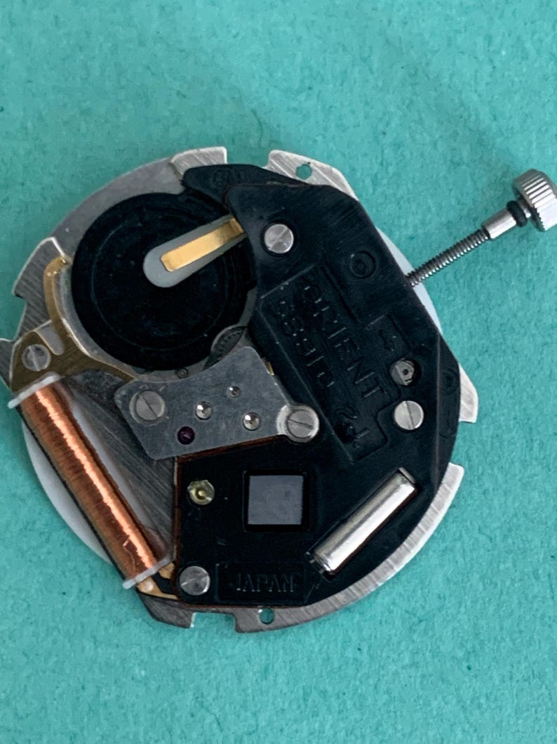 May include: Close-up of a watch movement with a black and silver design. The watch mechanism includes a battery, a copper coil, and the word "ORIENT". The watch movement is labeled "JAPAN".