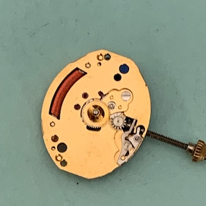 May include: A gold-colored watch movement with intricate gears, a red curved component, and a blue dot. A screw extends from the side. The watch mechanism is set against a teal background.