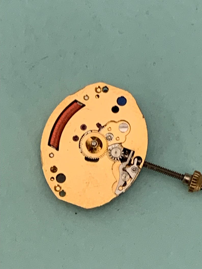 May include: A gold-colored watch movement with intricate gears, a red curved component, and a blue dot. A screw extends from the side. The watch mechanism is set against a teal background.