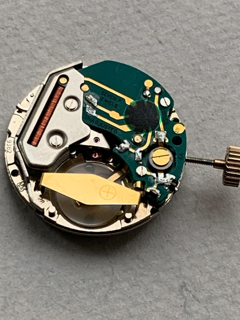 Puede incluir: Primer plano de un movimiento de reloj con un rotor dorado y una placa de circuito verde. La placa tiene el texto "SEVEN JEWELS" y "UNADJUSTED". El movimiento es circular con una corona dorada.