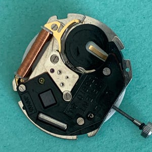 May include: A detailed view of a watch movement, showcasing its intricate parts. It includes a copper coil, gold-toned elements, and a black plate marked "ORIENT 55010 2J" and "JAPAN". A small silver screw is also present.