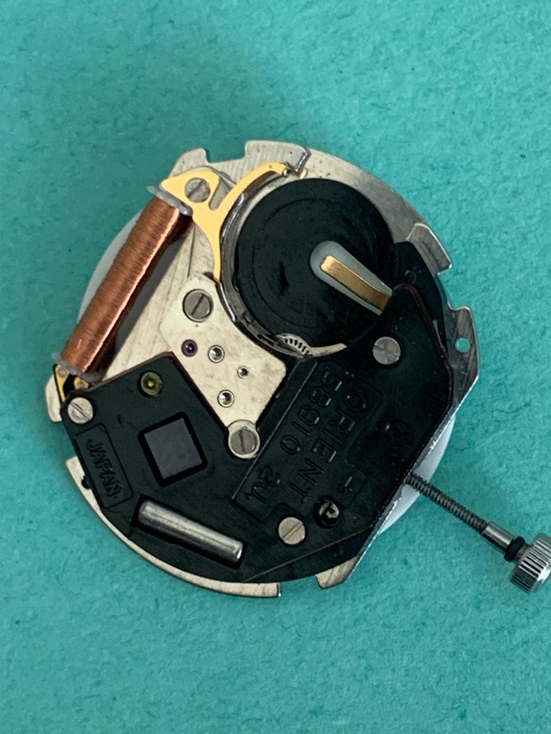 May include: A detailed view of a watch movement, showcasing its intricate parts. It includes a copper coil, gold-toned elements, and a black plate marked "ORIENT 55010 2J" and "JAPAN". A small silver screw is also present.