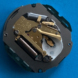 May include: Detailed view of a watch mechanism, revealing its internal workings. The image displays gold-toned gears, springs, and other elements, housed within a grey and green casing. The text "2110A" is printed on a component.