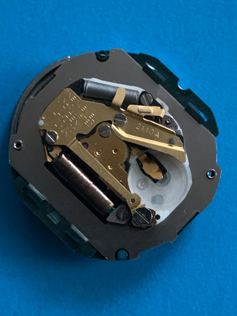 May include: Detailed view of a watch mechanism, revealing its internal workings. The image displays gold-toned gears, springs, and other elements, housed within a grey and green casing. The text "2110A" is printed on a component.