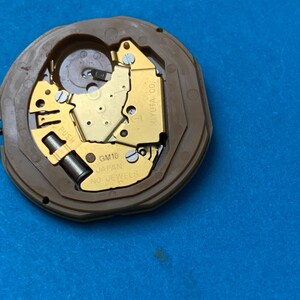 May include: A watch movement with a brown plastic casing and gold-colored internal components. The text "GM10 JAPAN NO JEWELS" is visible. The movement is set against a blue background.