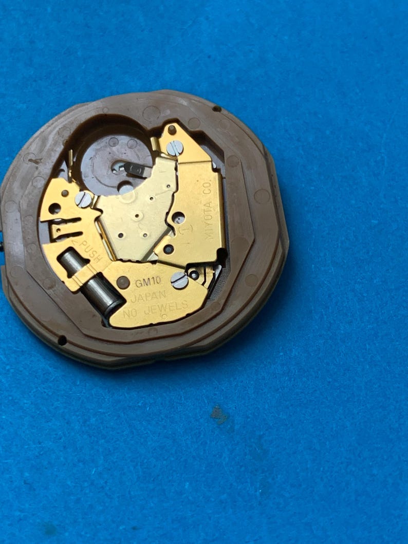 May include: A watch movement with a brown plastic casing and gold-colored internal components. The text "GM10 JAPAN NO JEWELS" is visible. The movement is set against a blue background.
