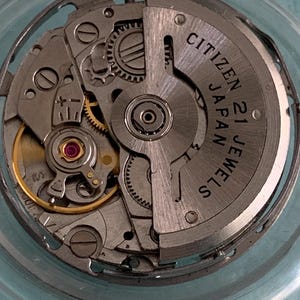 May include: Close-up of a Citizen 21 Jewels watch movement. The silver-toned mechanism features intricate gears, a red jewel, and the text "CITIZEN 21 JAPAN JEWELS". The watch movement is encased in a clear, circular frame.