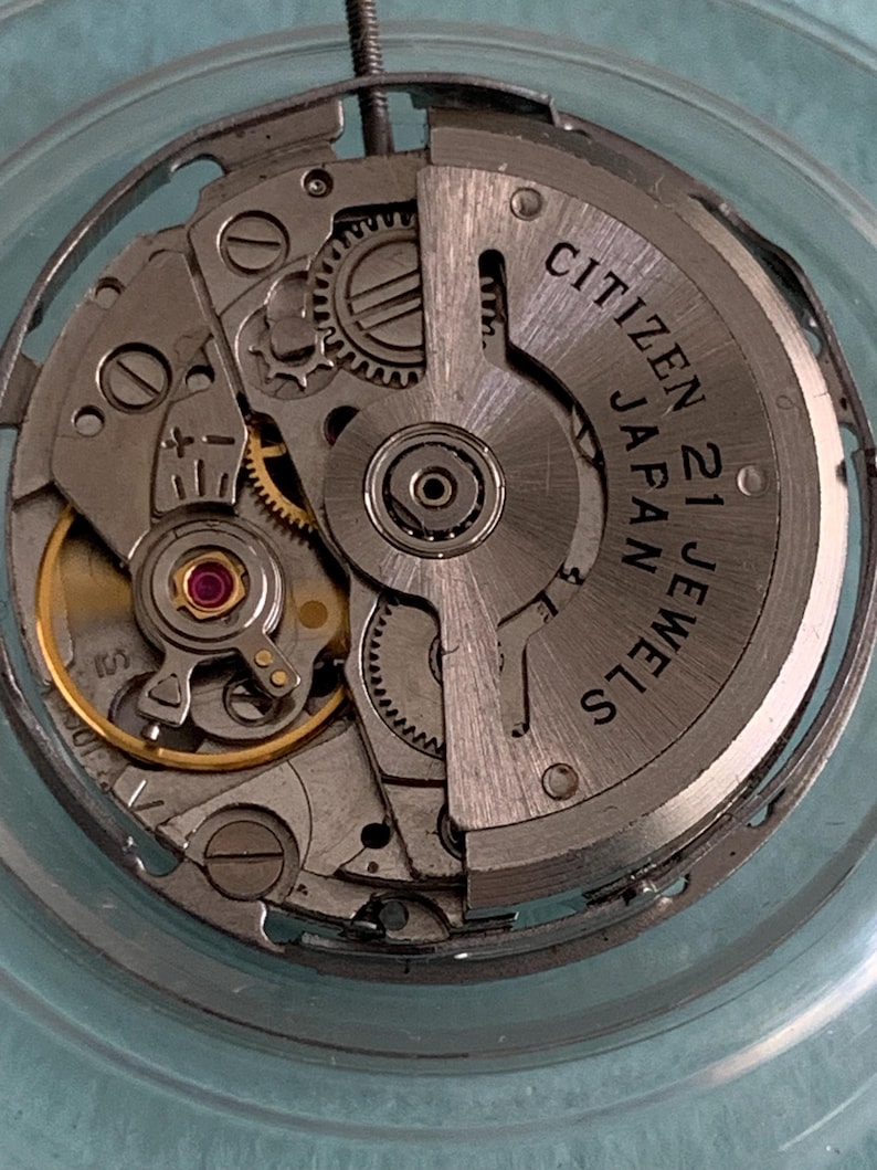 May include: Close-up of a Citizen 21 Jewels watch movement. The silver-toned mechanism features intricate gears, a red jewel, and the text "CITIZEN 21 JAPAN JEWELS". The watch movement is encased in a clear, circular frame.