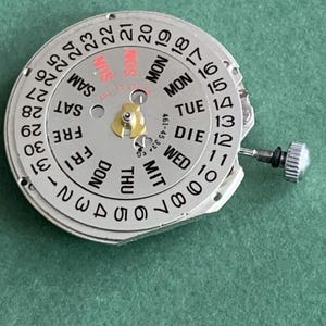 May include: A silver watch movement with a white dial showing the days of the week and numbered dates. The dial has a small gold-coloured component in the centre and a small silver knob on the side. The background is a solid green colour.