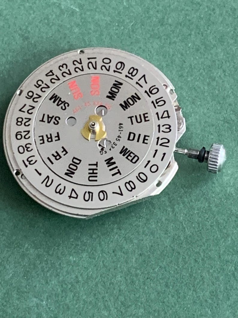 May include: A silver watch movement with a white dial showing the days of the week and numbered dates. The dial has a small gold-coloured component in the centre and a small silver knob on the side. The background is a solid green colour.