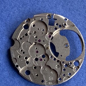 Puede incluir: Un movimiento de reloj plateado con intrincados engranajes, agujeros y joyas de rub&iacute;. El mecanismo circular tiene una gran abertura y varios componentes circulares m&aacute;s peque&ntilde;os. El n&uacute;mero "1695" est&aacute; grabado en el metal. El fondo es azul liso.