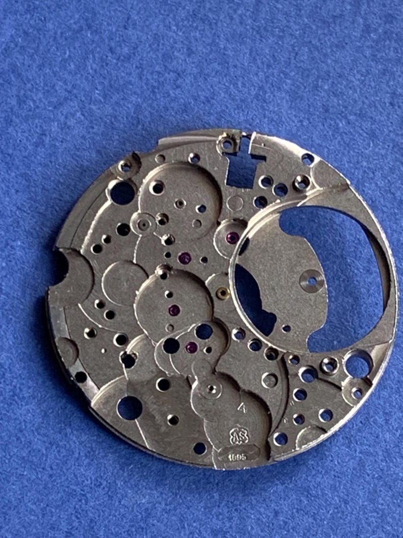 Puede incluir: Un movimiento de reloj plateado con intrincados engranajes, agujeros y joyas de rub&iacute;. El mecanismo circular tiene una gran abertura y varios componentes circulares m&aacute;s peque&ntilde;os. El n&uacute;mero "1695" est&aacute; grabado en el metal. El fondo es azul liso.