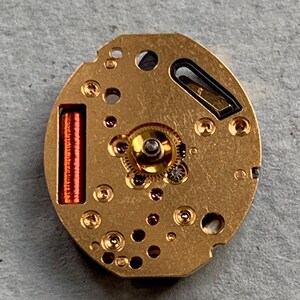 May include: A detailed view of a gold-coloured watch movement, showcasing tiny gears, ruby jewels, and a red coil. The oval mechanism has several holes and a small black element. The watch component is set against a grey background.