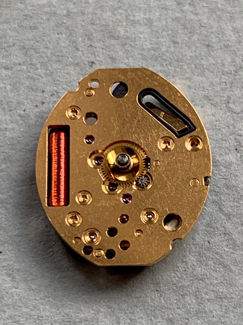 May include: A detailed view of a gold-coloured watch movement, showcasing tiny gears, ruby jewels, and a red coil. The oval mechanism has several holes and a small black element. The watch component is set against a grey background.