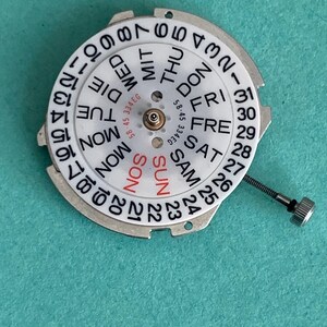 May include: A watch movement component with a white dial featuring black numbers and day abbreviations. The days of the week are in a circle, with "SUN" in red. A small metal screw is attached. The component is on a teal surface.