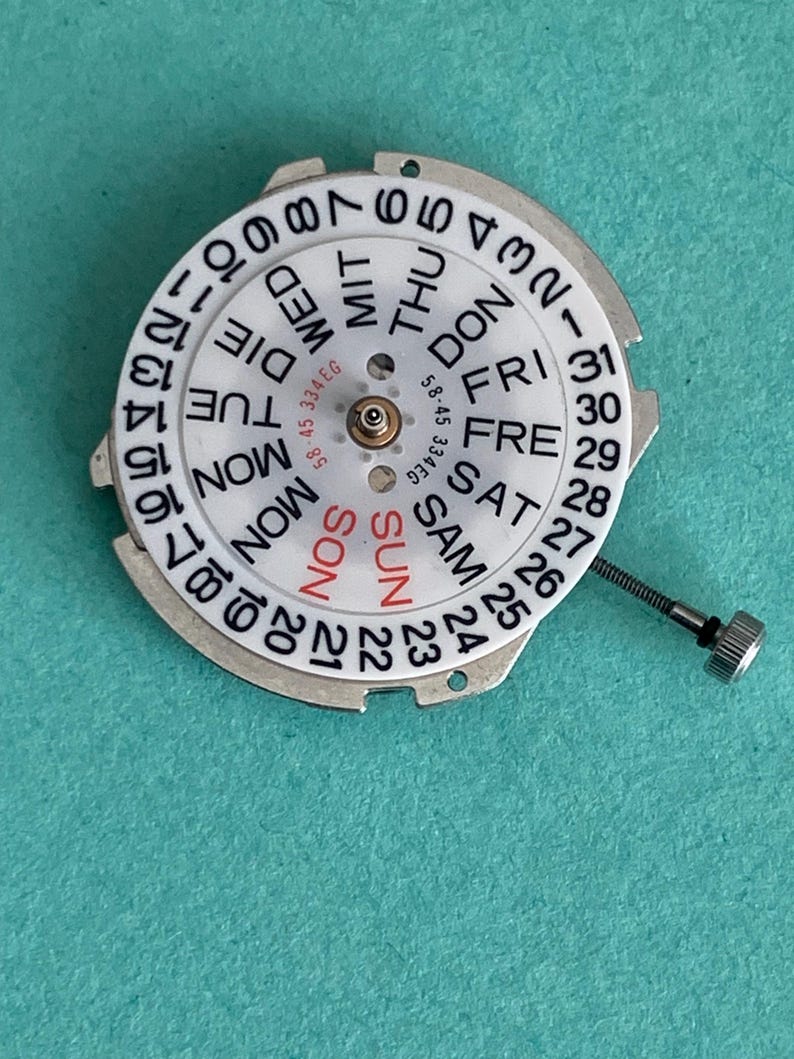 May include: A watch movement component with a white dial featuring black numbers and day abbreviations. The days of the week are in a circle, with "SUN" in red. A small metal screw is attached. The component is on a teal surface.