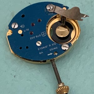 May include: A close-up of a watch movement with a blue circuit board and gold-colored components. The watch movement has the text "SWISS SIX 6 JEWELS" and "978.002" printed on it. A gold-colored crown is attached to the movement.
