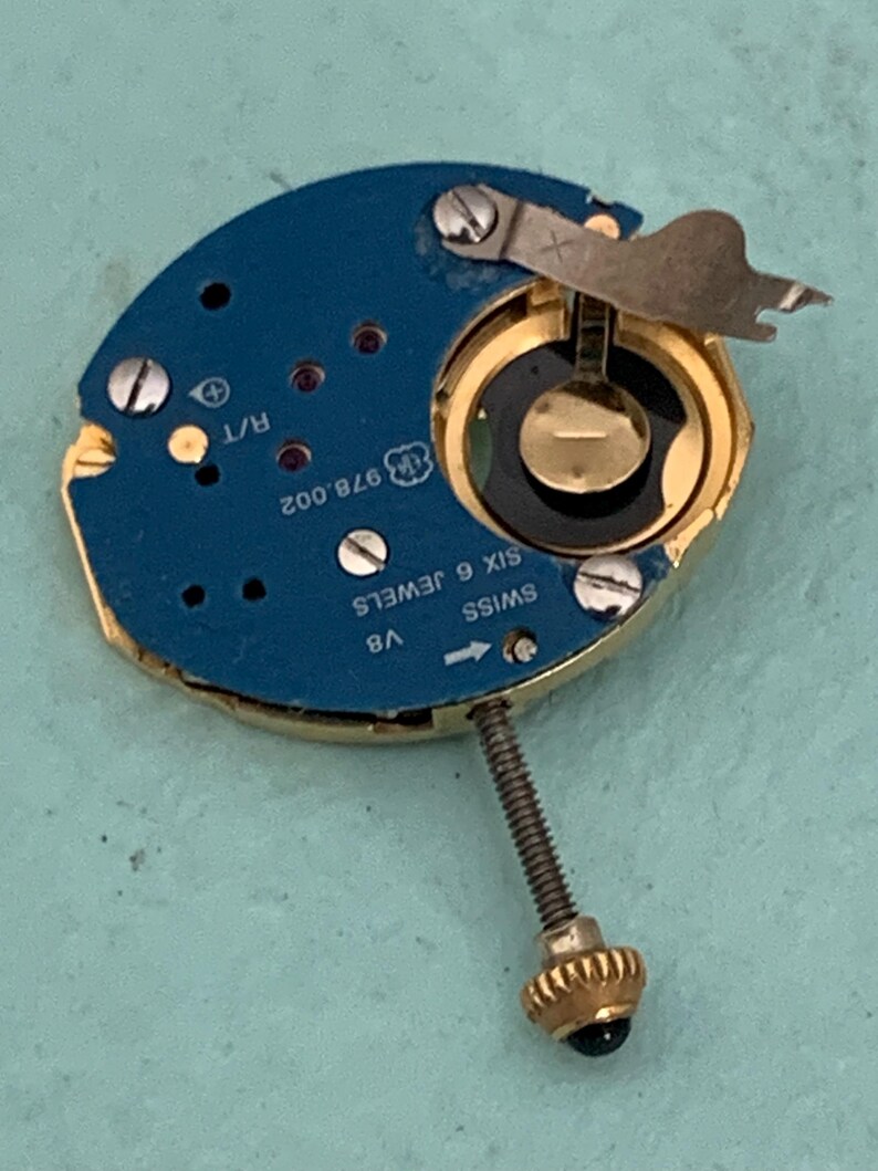 May include: A close-up of a watch movement with a blue circuit board and gold-colored components. The watch movement has the text "SWISS SIX 6 JEWELS" and "978.002" printed on it. A gold-colored crown is attached to the movement.