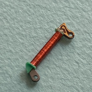 May include: A small, copper-colored coil with a green ring and two metal end pieces. One end piece is a rounded shape with a hole, and the other is a decorative shape with a hole. The coil is on a light blue surface.