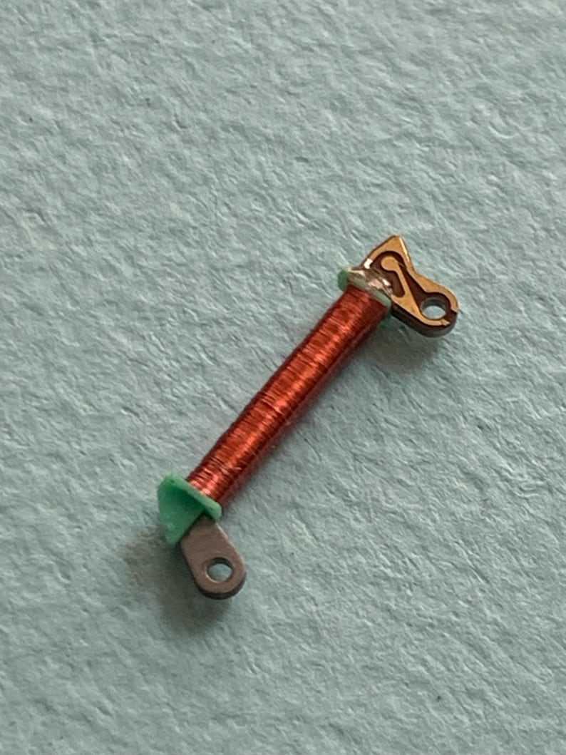 May include: A small, copper-colored coil with a green ring and two metal end pieces. One end piece is a rounded shape with a hole, and the other is a decorative shape with a hole. The coil is on a light blue surface.