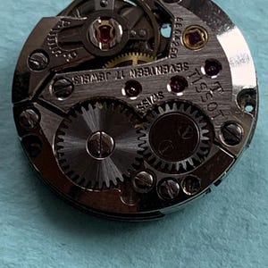 May include: A silver Tissot watch movement with the text "Tissot" and "Seventeen Jewels" printed on the face. The movement has a silver gear system and a red jewel.