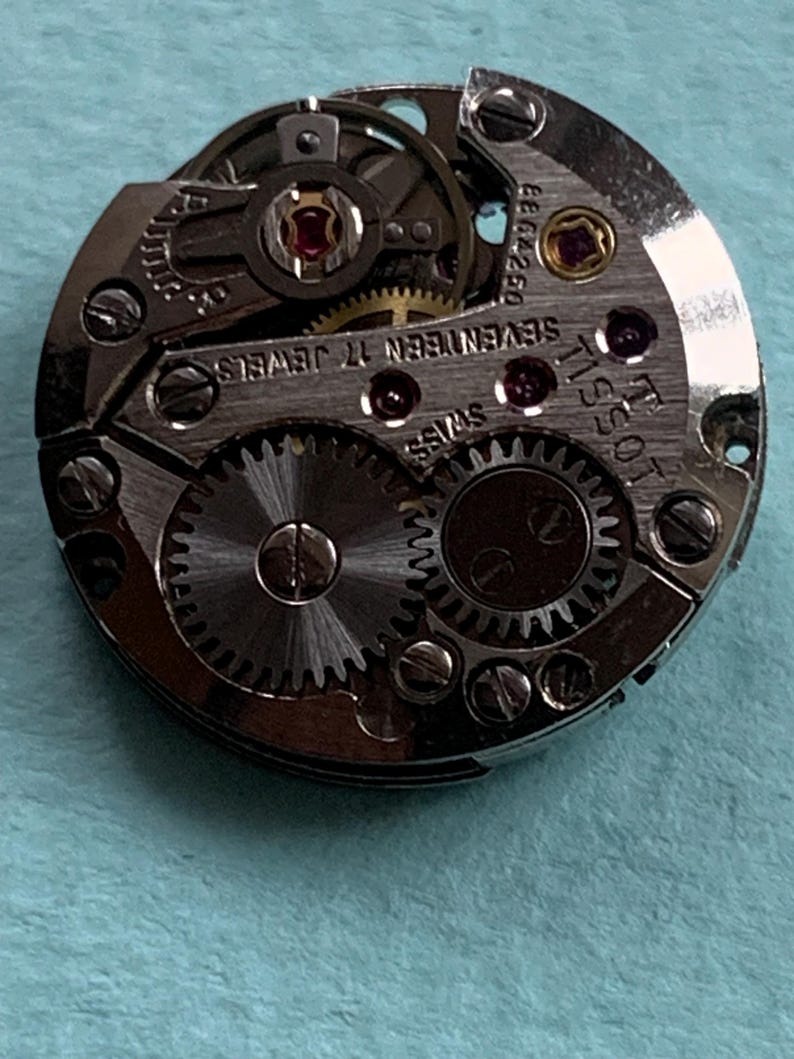 May include: A silver Tissot watch movement with the text "Tissot" and "Seventeen Jewels" printed on the face. The movement has a silver gear system and a red jewel.