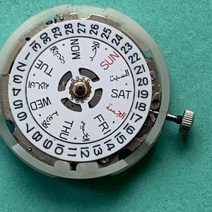 May include: A close-up of a watch movement with a white dial and Arabic numerals. The dial displays the days of the week in English and Arabic, along with the date. A small crown is visible on the side. The background is a teal color.