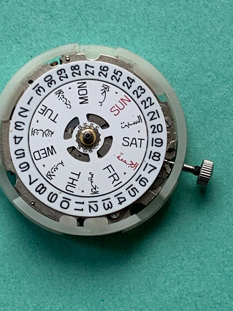 May include: A close-up of a watch movement with a white dial and Arabic numerals. The dial displays the days of the week in English and Arabic, along with the date. A small crown is visible on the side. The background is a teal color.