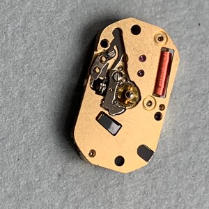 May include: Close-up of a gold-colored watch movement with intricate gears, a red coil, and small black and purple components. The rectangular piece has several holes and is set against a gray surface.