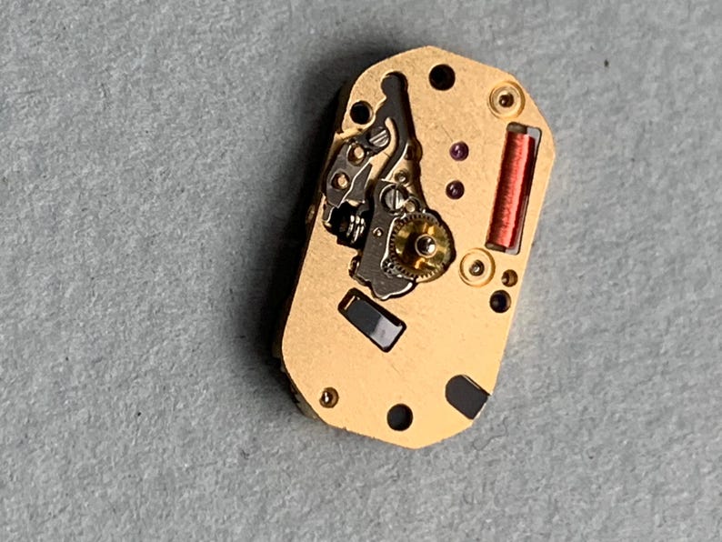 May include: Close-up of a gold-colored watch movement with intricate gears, a red coil, and small black and purple components. The rectangular piece has several holes and is set against a gray surface.