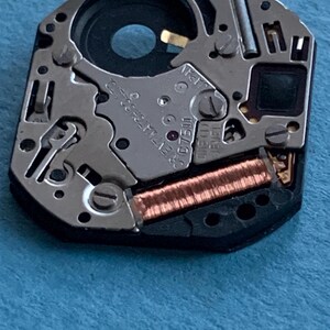 May include: Close-up of a watch movement with intricate mechanical components. The movement features silver-colored metal parts, a copper-colored coil, and the text "JAPAN". The watch mechanism is set against a blue background.