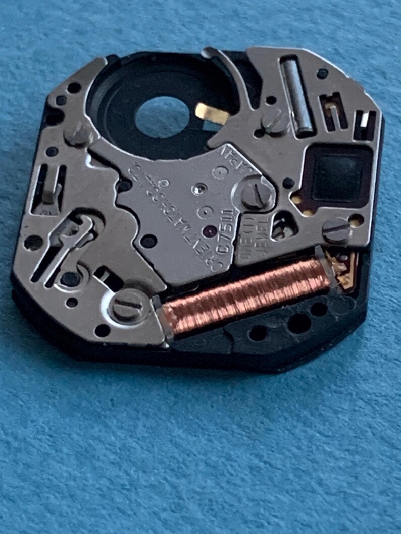 May include: Close-up of a watch movement with intricate mechanical components. The movement features silver-colored metal parts, a copper-colored coil, and the text "JAPAN". The watch mechanism is set against a blue background.