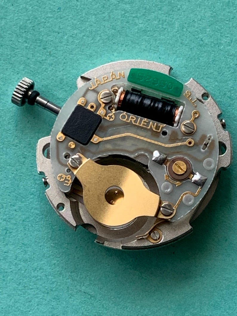 May include: A close-up of a watch movement with the word "JAPAN" and "ORIENT" visible. The movement has a silver-colored base with gold and black components, including a black coil and a green piece with numbers.